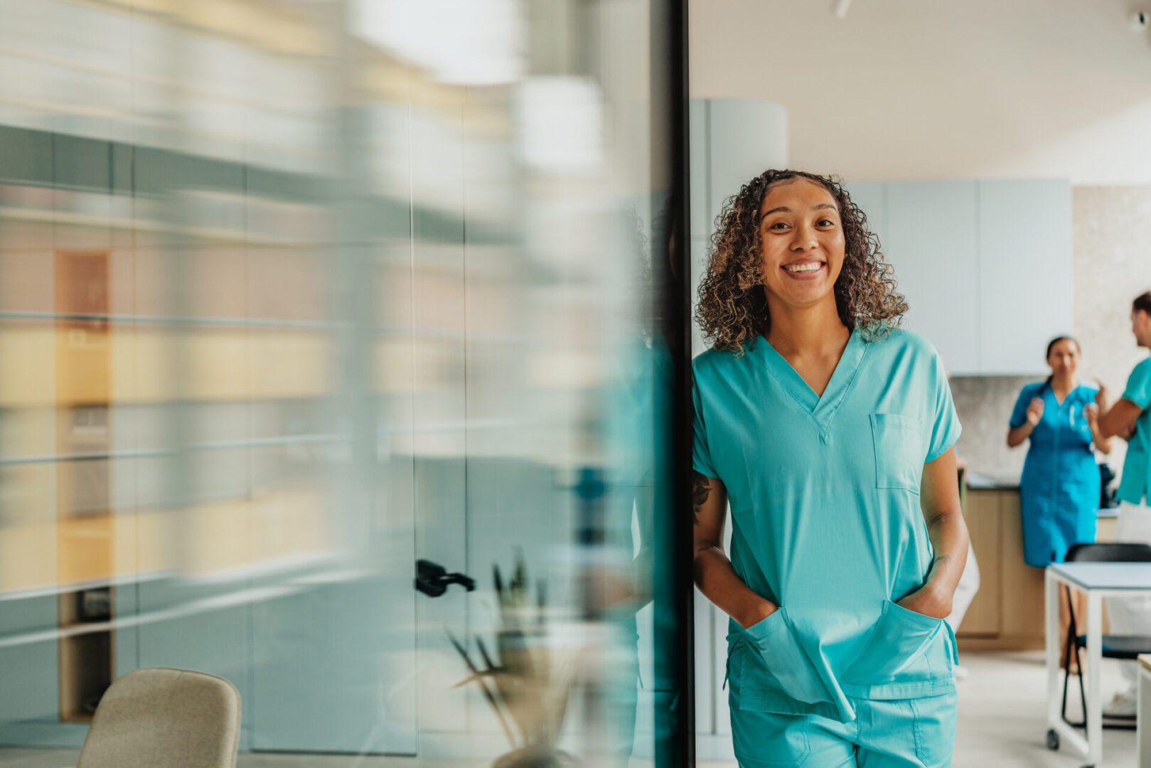 Smiling nurse in scrubs, colleagues in background.